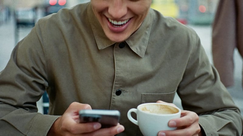 Man smiling at his phone after a new Hinge match, sitting outdoors with coffee