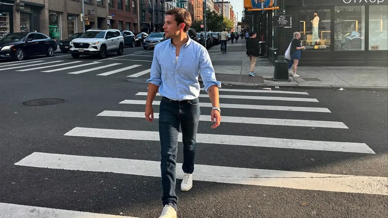 man in a light blue shirt and jeans walking across a city crosswalk in afternoon sun as a full body dating photo example
